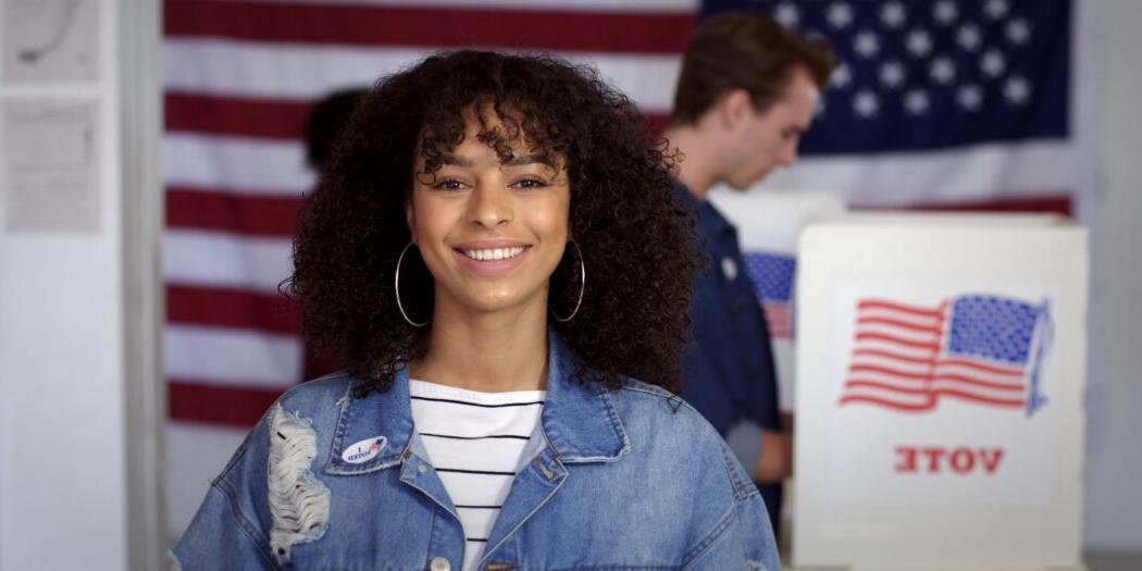 Woman wearing a jean jacket and white striped shirt standing in front of a voting booth smiling with the American flag behind her 